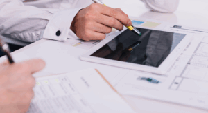 Profissionais analisando projetos técnicos e plantas arquitetônicas em uma mesa, utilizando um tablet e canetas para revisar detalhes de construção.