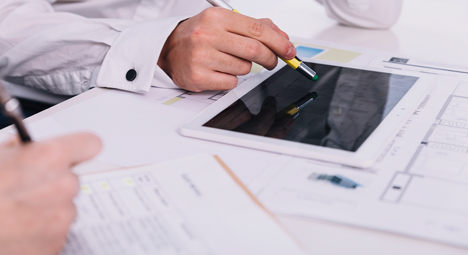 Profissionais analisando projetos técnicos e plantas arquitetônicas em uma mesa, utilizando um tablet e canetas para revisar detalhes de construção.