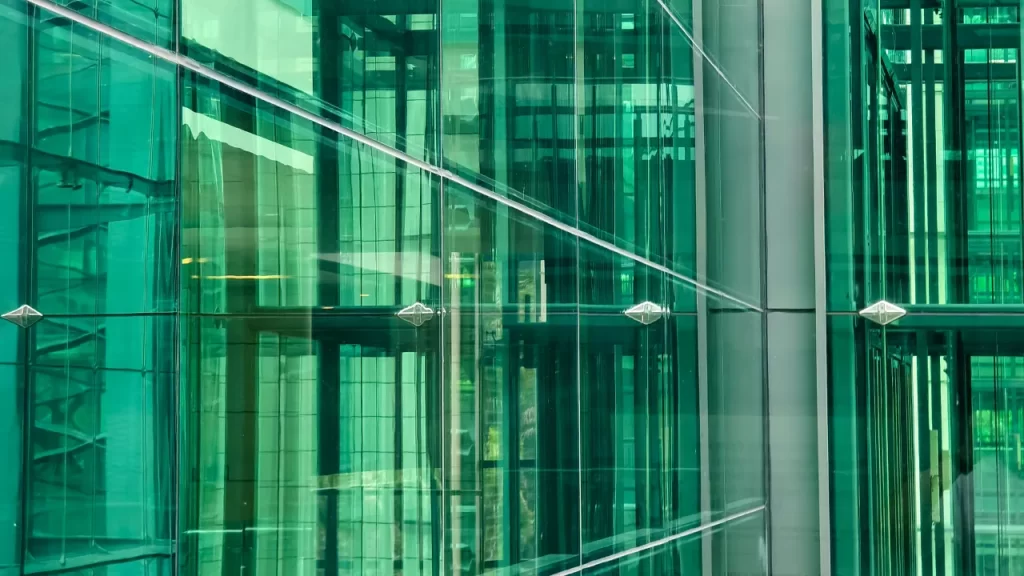 Fachada moderna de edifício composta por painéis de vidro verde, com estrutura metálica refletindo elementos internos e externos.