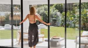 Mulher de costas abrindo uma porta de vidro deslizante, com vista para um jardim amplo e iluminado, cercado por plantas e área externa decorada.