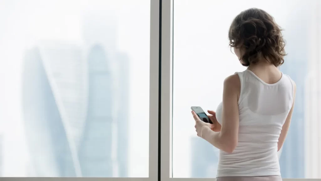 Mulher de costas segurando um smartphone enquanto olha pela janela para prédios altos desfocados.
